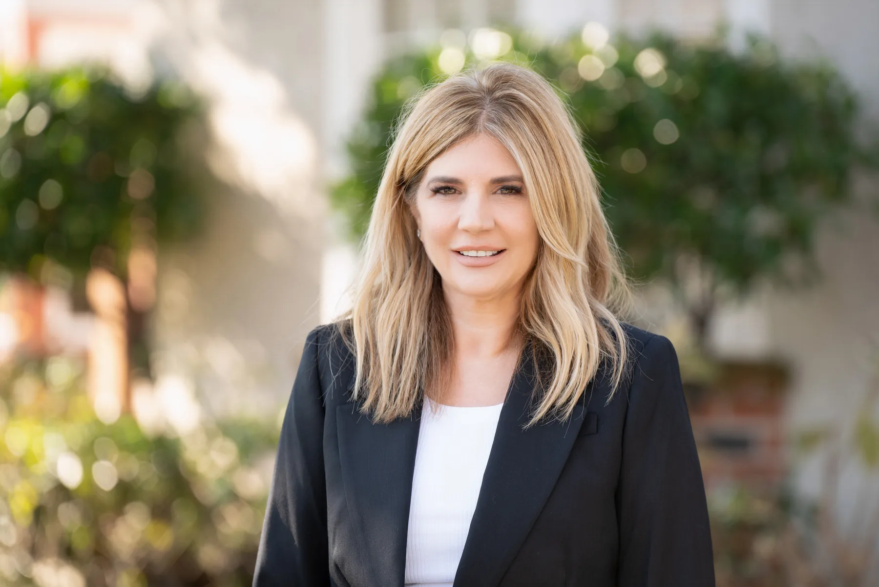 Michele Kesner outdoors under trees, smiling, in a black blazer.